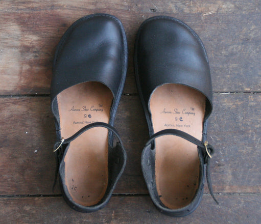 A worn pair of black Mary Jane shoes by Aurora Shoe Co. top down on an old, worn wooden floor.   The wear of the insole and the soft texture of the leather are apparent in the soft light. 