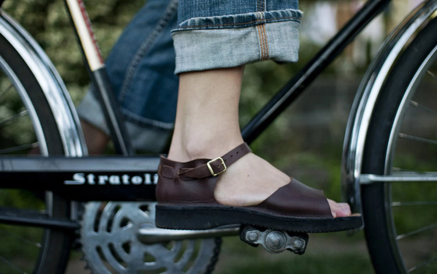 Model is wearing a brown pair of Handmade Leather New Mexican Sandals by Aurora Shoe Co. The model is wearing rolled blue jeans and no socks, pedaling a vintage black bicycle with chrome fenders.