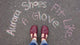 A customer is wearing a pair of Burgundy Middle T-Strap shoes by Aurora Shoe Co. The photo is taken from the customer's perspective, looking down at her new burgundy handmade T-straps. In Chalk, the words "Aurora Shoes Fit Like A Glove" is written on a blacktop surface, framing her new shoes in the photo.