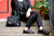 Woman sitting on steps with red door and plants in background, next to black wrought iron railing, wearing black top, black pants, sporting a black leather handbag and black Aurora Shoe Co. Middle English Handmade Leather Shoes.