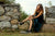 Model wearing Aurora Shoe Co. T-Strap handmade leather shoes in black, sitting on stone wall, wearing dress.