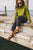 Woman sitting on dock at Cayuga Lake, wearing sunglasses, a green shirt, black pants, and Olive Aurora Shoe Co. T-Strap leather shoes.
