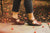 Woman walking in fall, on sidewalk, with fallen flowers, wearing black cutoff jeans and Brown Aurora Shoe Co. Handmade T-strap shoes.