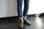 Legs of model, wearing blue jeans, rolled cuff, on black floor adjacent to white wall, wearing Aurora Shoe Co. Olive Middle English Leather Shoes.