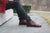 Woman sitting on stone steps at Wells College wearing red jacket, black pants, printed socks, and burgundy T-strap Aurora Shoe Co. Handmade Leather Shoes - side view.