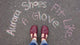 A customer is wearing a pair of Burgundy Middle T-Strap shoes by Aurora Shoe Co.    The photo is taken from the customer's perspective, looking down at her new burgundy handmade T-straps.   In Chalk, the words "Aurora Shoes Fit Like A Glove" is written on a blacktop surface, framing her new shoes in the photo.