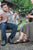 Couple sitting in adirondack chairs, playing guitar around campfire, wearing Aurora Shoe Co. Handmade Leather Shoes.   Man is wearing Men's Middle English, Navy.   Woman is wearing Women's New Mexican sandal in Olive color.  In this orientation, the camera is focused on the man, with a beard, wearing glasses, and playing an acoustic guitar.  