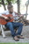 Man with beard and glasses playing guitar with lake in background, sitting on wooden adirondack chair, wearing Men's Middle English, Olive Handmade Leather Shoes by Aurora Shoe Co.