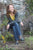 woman sitting in front of stone wall, smiling, plants around, wearing yellow shirt, blue jeans, and Navy T-strap handmade leather shoes by Aurora Shoe Co.