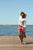 woman standing on wood dock, holding sun hat, in front of Cayuga Lake at Aurora, New York, holding burgundy New Mexican handmade leather shoes in one hand.