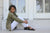 Side view of Woman sitting on stone steps, wearing green jacket, white pants, and black New Mexican handmade leather sandals by Aurora Shoe Co.