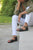 Second view of Woman sitting on stone steps, wearing green jacket, white pants, and black New Mexican handmade leather sandals by Aurora Shoe Co.