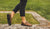 Model sanding on stone wall, wearing black cutoff jeans, in fall with grass in background, advertising Aurora Shoe Co. Women's Middle English Olive Handmade Leather shoes.