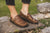 Model standing on flat stone with green plans around, wearing Aurora Shoe Co. Olive Handmade  Leather Middle English Shoes.